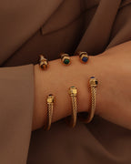 Tres pulseras rígidas abiertas doradas con textura de cable y extremos de colores verde, azul y marrón, puestas en la muñeca.