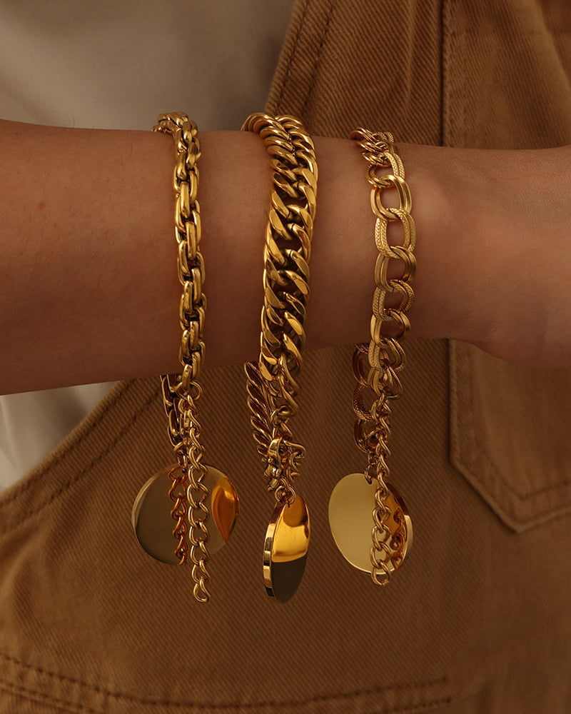 Tres pulseras doradas de cadenas de distintos eslabones con discos redondos colgantes, puestas en la muñeca.