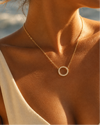 Persona con collar fino dorado y colgante circular abierto de pequeñas piedras claras, con luz de sol sobre la piel.