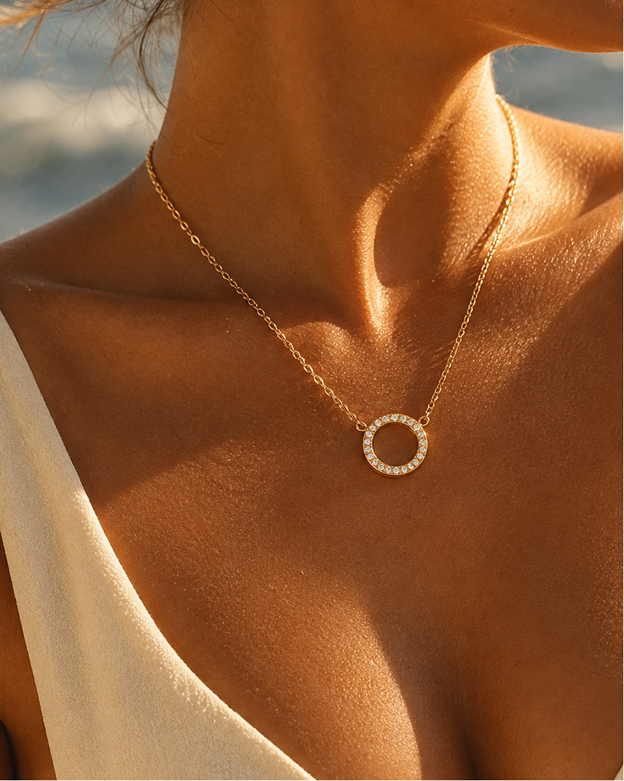 Persona con collar fino dorado y colgante circular abierto de pequeñas piedras claras, con luz de sol sobre la piel.