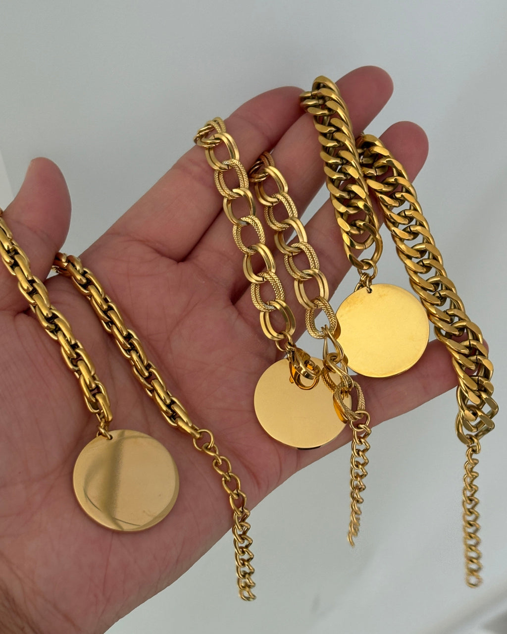 Tres pulseras doradas de cadenas de distintos grosores con discos redondos colgantes, sostenidas en una mano.