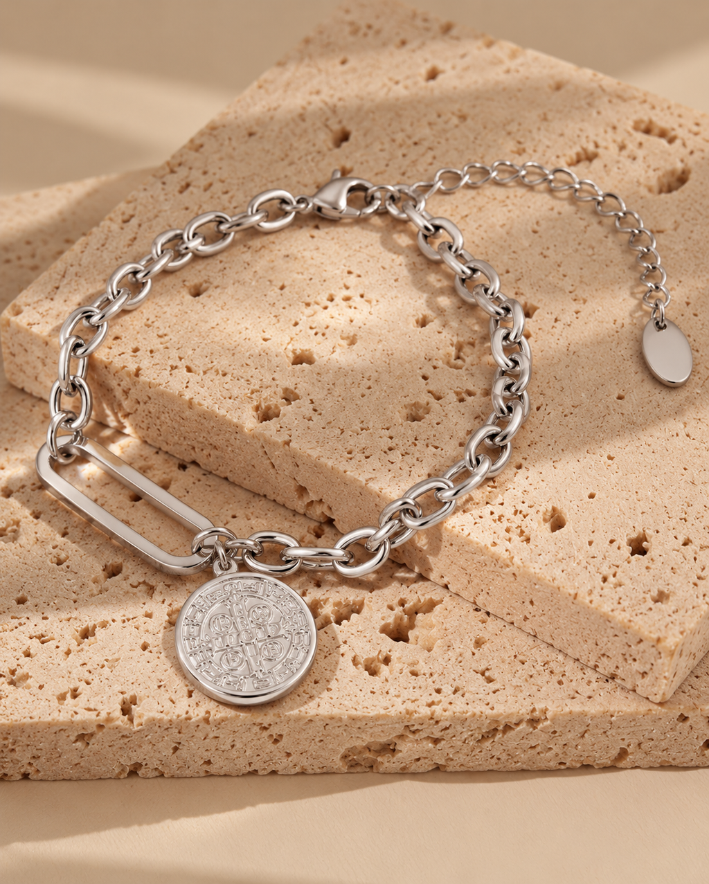 Pulsera de tono plateado con eslabones ovalados, eslabón central alargado y medalla redonda grabada, sobre piedra beige.