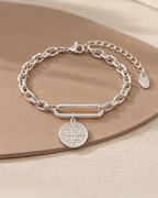 Pulsera de tono plateado con eslabón central alargado, medalla redonda grabada y cadena de extensión, sobre superficie beige.