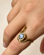 Primer plano de un anillo dorado de Xihe By Dm con diseño curvo abierto y piedra ovalada transparente, sobre fondo marfil.
