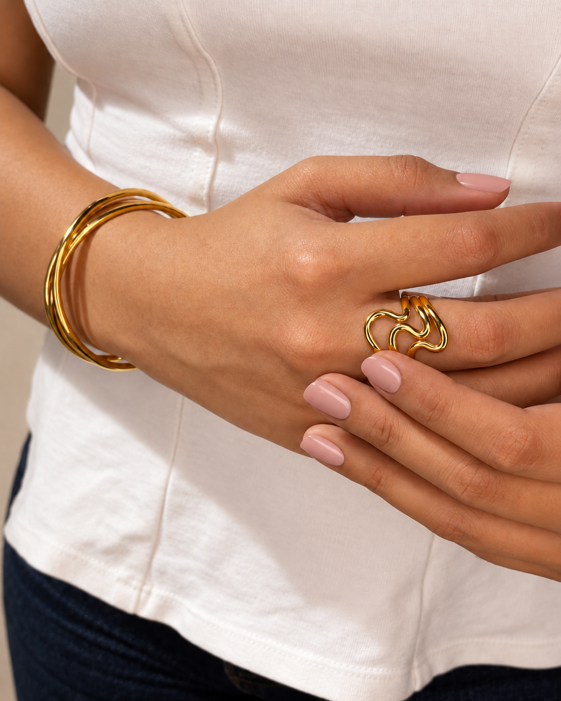 Manos con uñas nude luciendo un anillo dorado de ondas entrelazadas y pulseras rígidas doradas de Xihe By Dm sobre una blusa blanca.