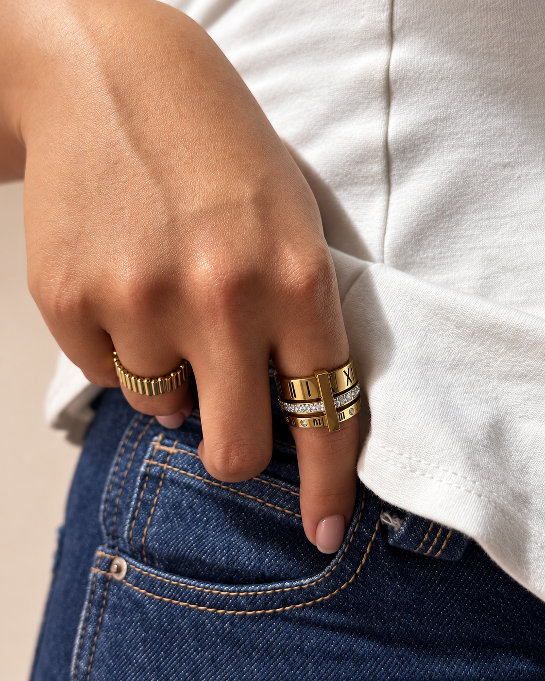 Mano con uñas nude luciendo un anillo dorado acanalado y otro de varias bandas con números romanos y piedras brillantes de Xihe By Dm, junto a un bolsillo de jeans.