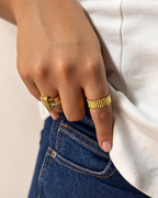 Mano con uñas nude luciendo dos anillos dorados de Xihe By Dm, uno con números romanos y piedras brillantes y otro acanalado, junto a jeans azules.