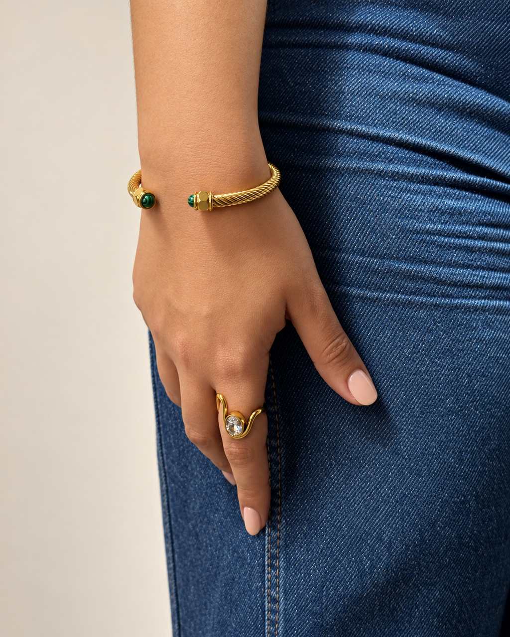 Mano con uñas nude, brazalete dorado trenzado con piedras verdes y anillo dorado con piedra ovalada brillante de Xihe By Dm, junto a jeans azules.