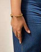 Mano con uñas nude, brazalete dorado trenzado con piedras verdes y anillo dorado con piedra ovalada brillante de Xihe By Dm, junto a jeans azules.