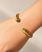 Primer plano de un brazalete abierto dorado de Xihe By Dm, con textura trenzada y piedras verdes en ambos extremos, sobre fondo marfil.