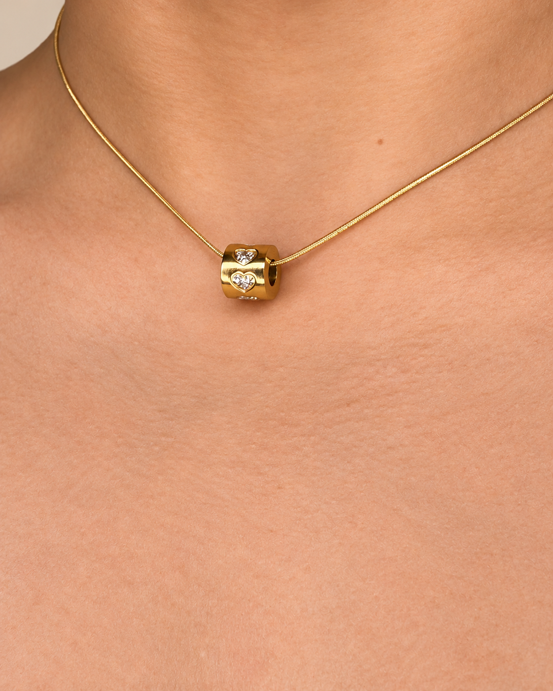 Primer plano de un collar dorado de Xihe By Dm con colgante cilíndrico pulido y detalles de corazones brillantes.