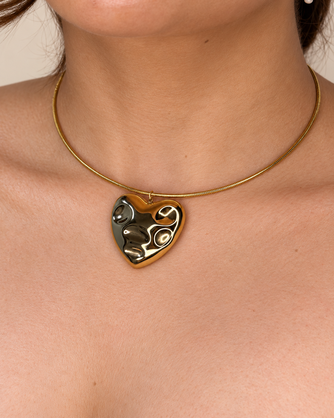 Collar dorado de Xihe By Dm con colgante de corazón de superficie ondulada y pulida, sobre el escote de una modelo.