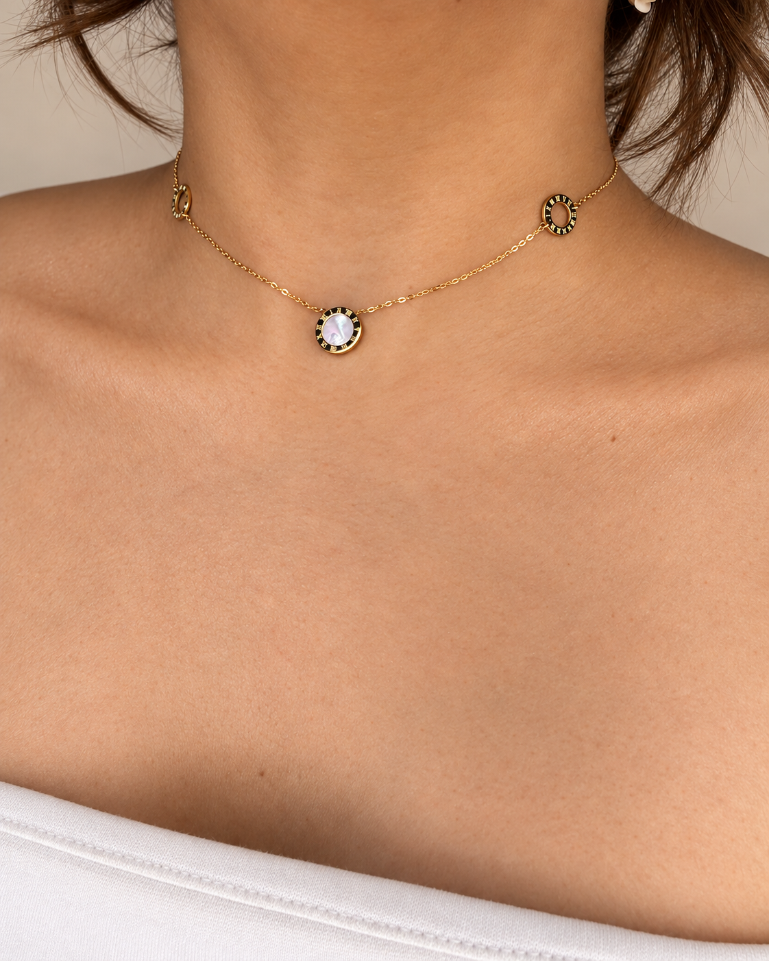 Collar dorado de Xihe By Dm con centro nacarado y aros con números romanos, lucido por una modelo con blusa blanca sobre fondo marfil.