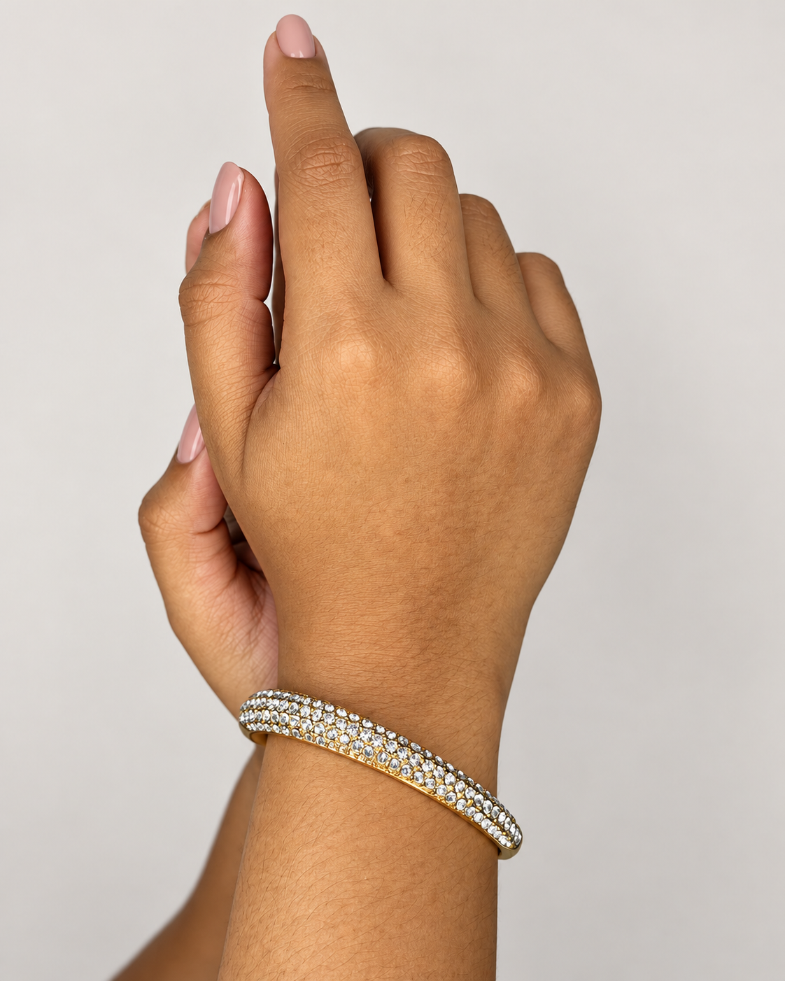 Pulsera rígida dorada con varias filas de piedras brillantes de Xihe By Dm, en la muñeca de una modelo con uñas nude sobre fondo claro.