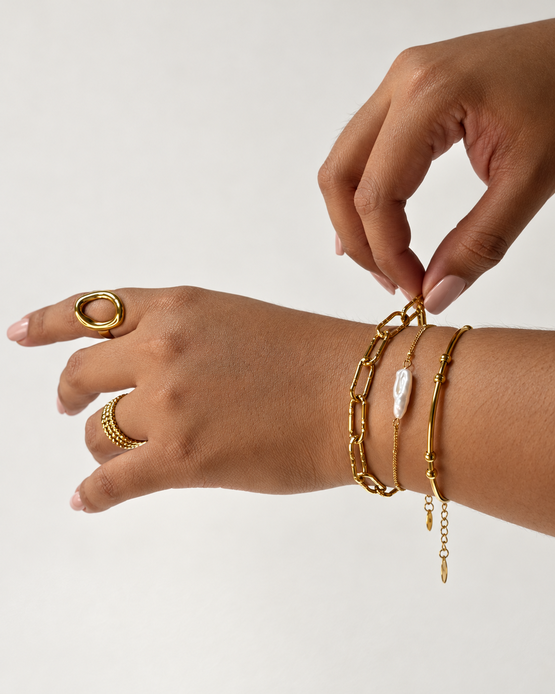 Manos con uñas nude luciendo anillos y tres pulseras doradas de Xihe By Dm, una de ellas con una perla blanca alargada.
