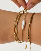 Primer plano de tres pulseras doradas de Xihe By Dm: una de eslabones, una cadena fina con perla blanca alargada y una pulsera tubular con cuentas.