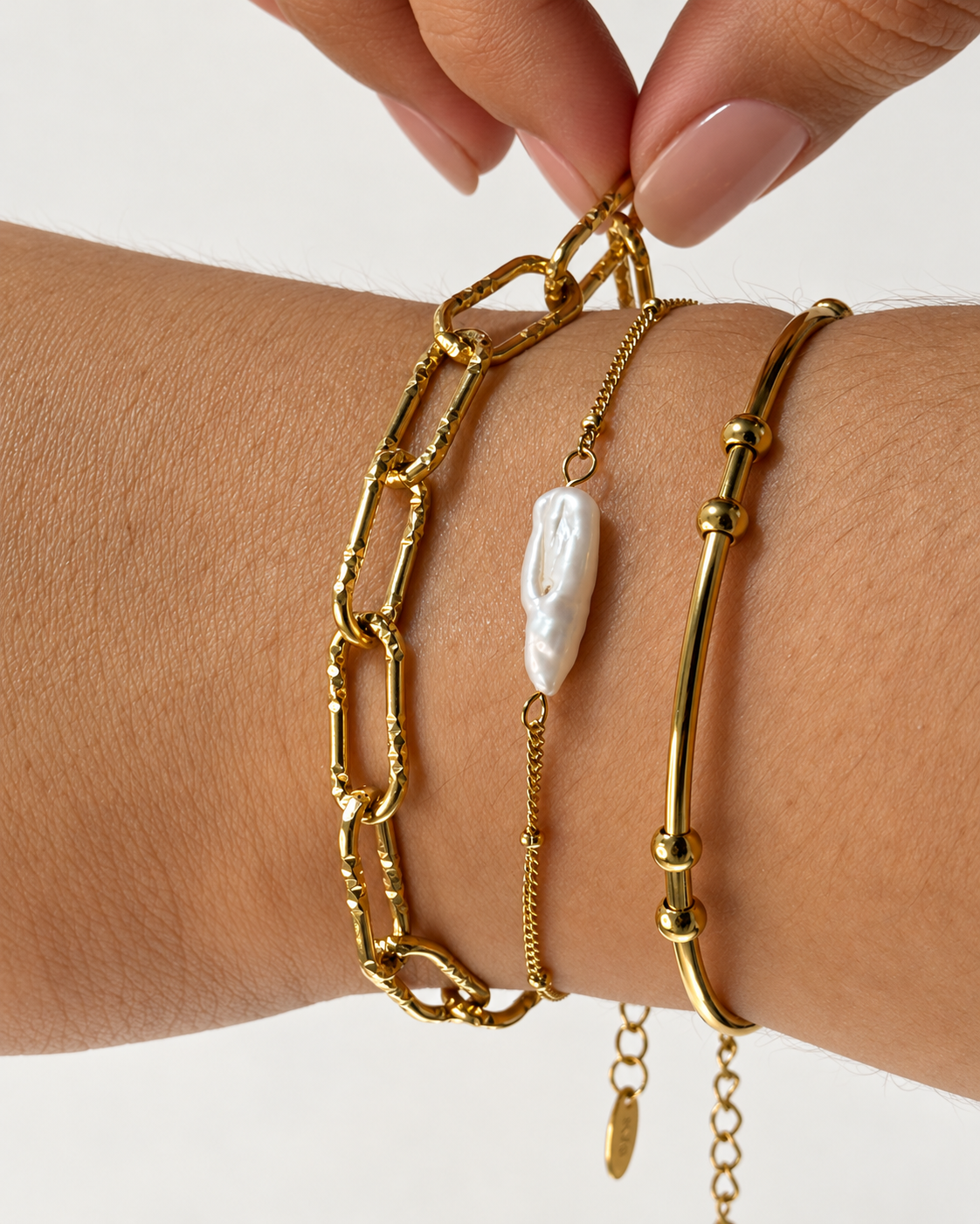 Primer plano de tres pulseras doradas de Xihe By Dm: una de eslabones, una cadena fina con perla blanca alargada y una pulsera tubular con cuentas.
