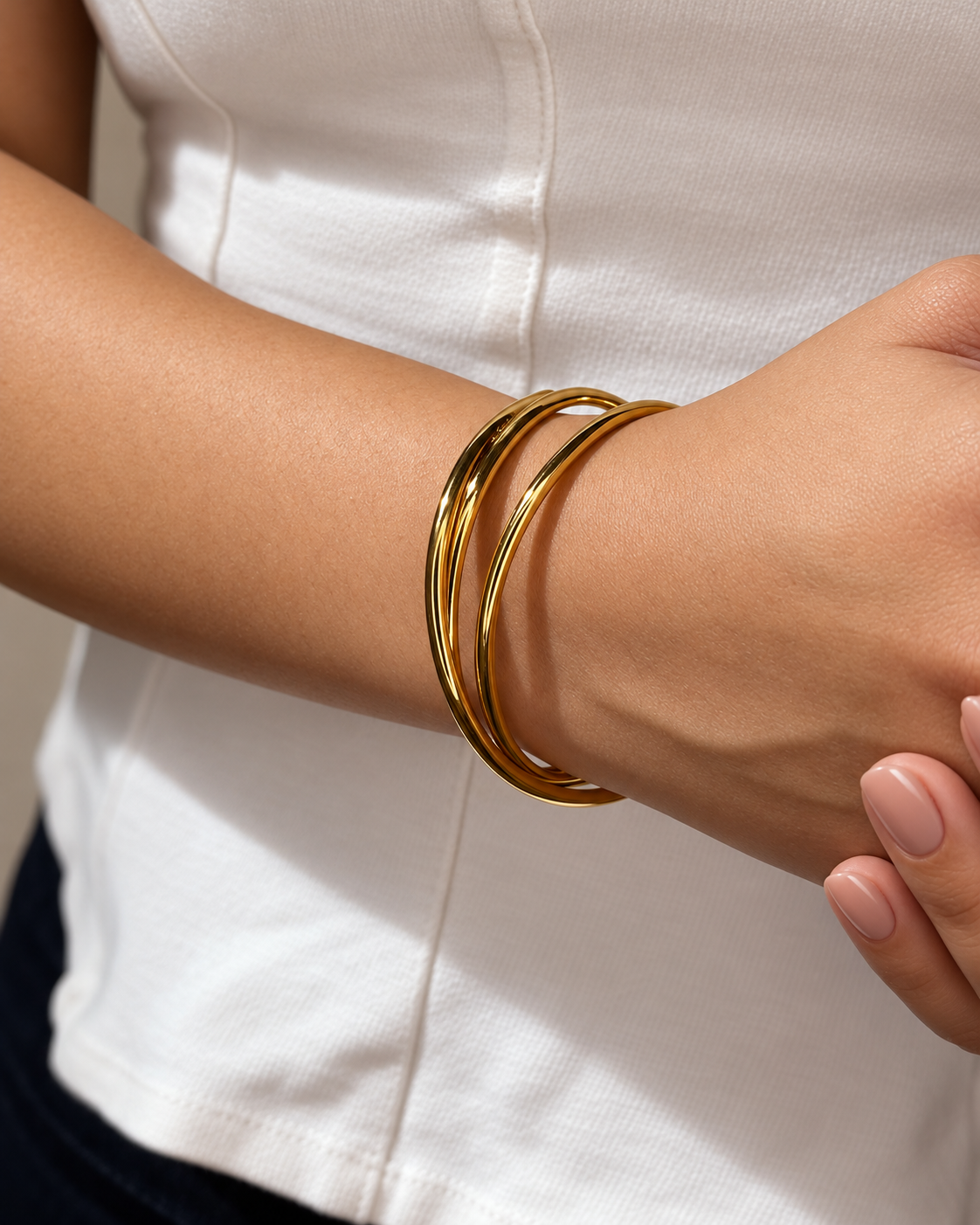 Primer plano de pulseras rígidas doradas de Xihe By Dm en la muñeca de una modelo con uñas nude, sobre una blusa blanca.
